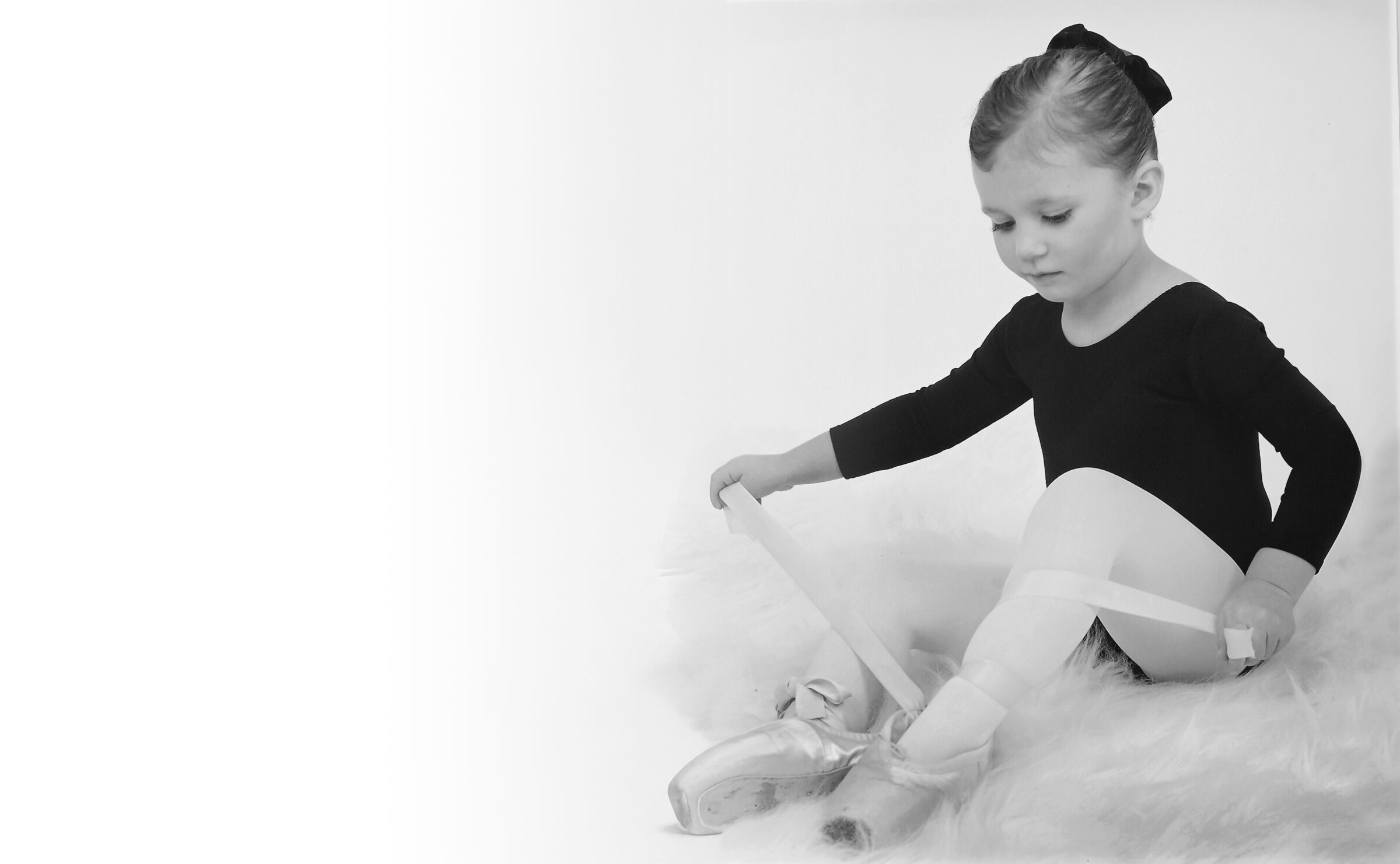 Young child in ballet attire sits on a fluffy surface, adjusting pointe shoes. Background is plain and bright. Black and white photograph.