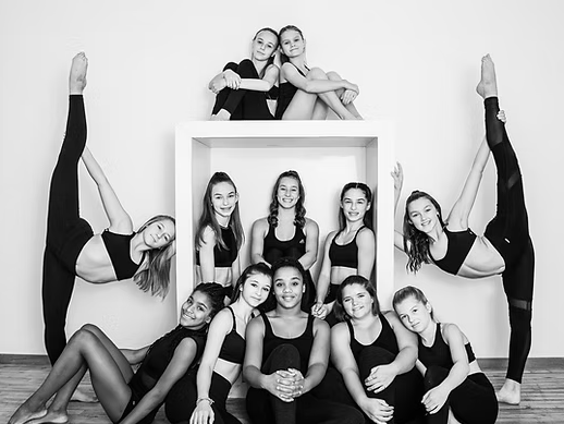 A group of young dancers poses creatively around a white cube in a dance studio, showcasing flexibility and teamwork in black outfits.