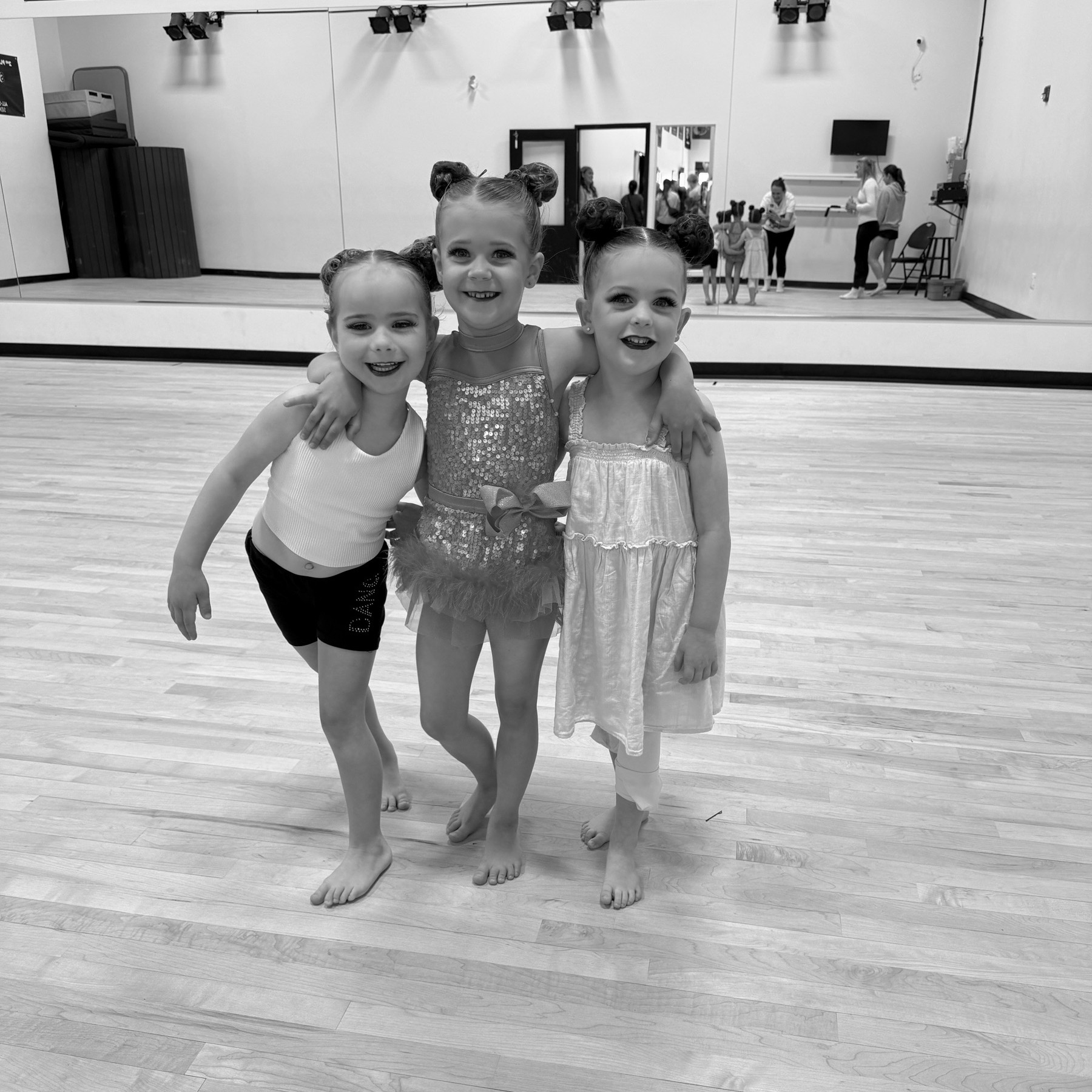 Three smiling children in dance attire pose in a studio with hardwood floors. Mirror reflects people in background.