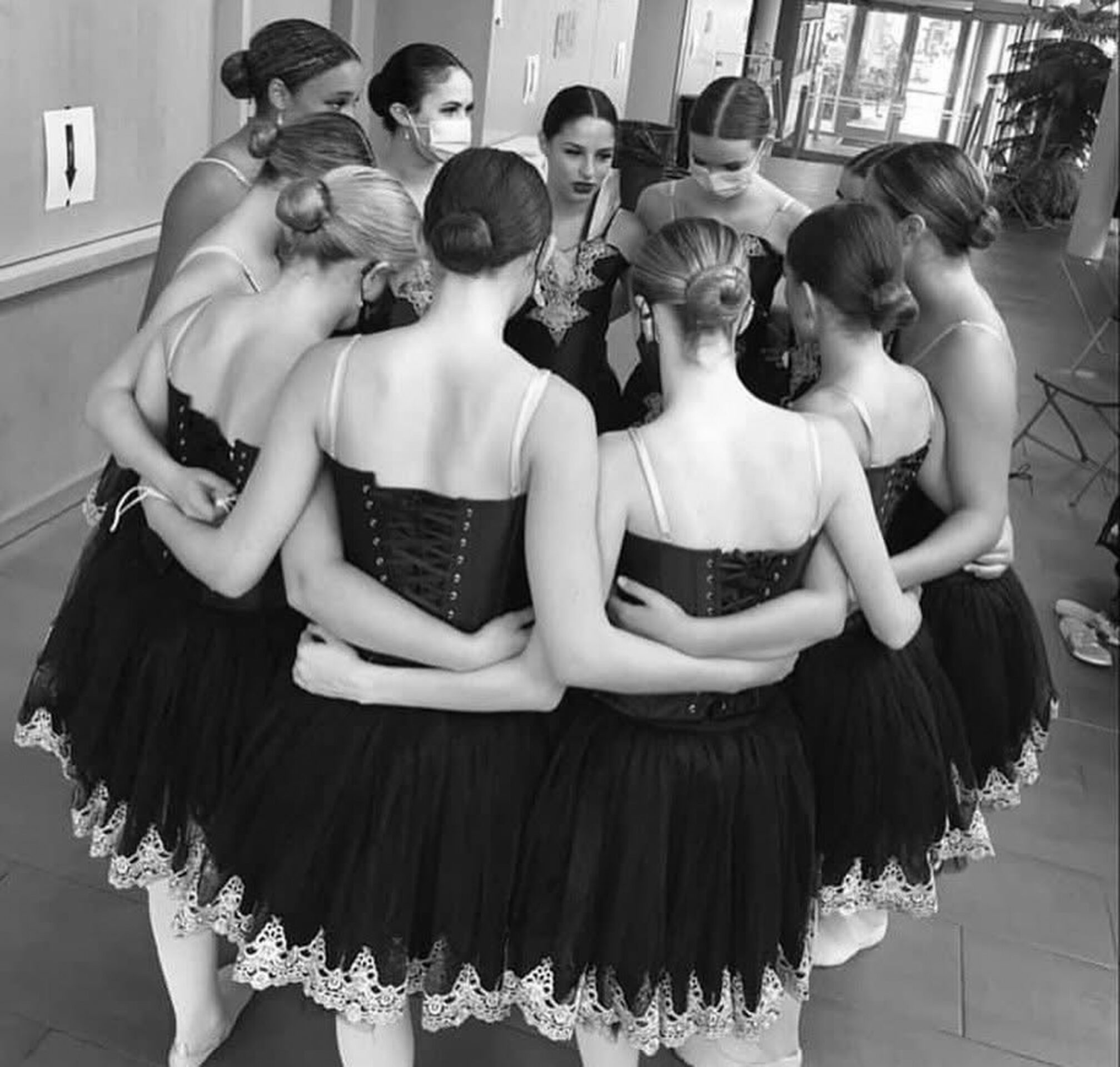 A group of eight ballet dancers huddle together, wearing elegant tutus and ballet shoes, possibly preparing for a performance in a backstage setting.