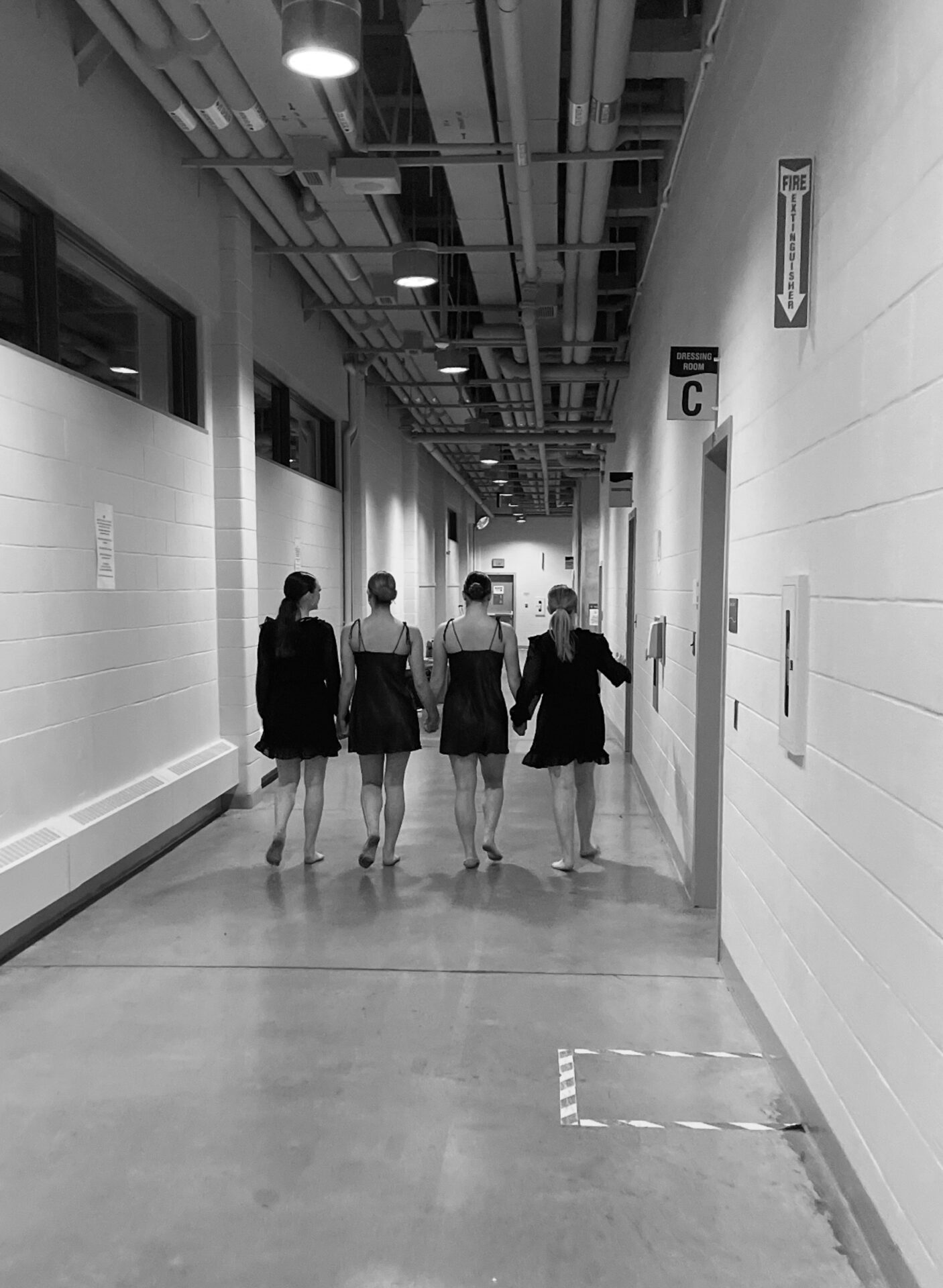 Four people in black dresses walk down a long, industrial corridor, indicating a backstage or rehearsal area. No landmarks or historical buildings visible.