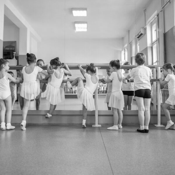 Children in ballet attire practice at a dance studio, using the barre for support. Mirrors reflect their movements, enhancing training focus.