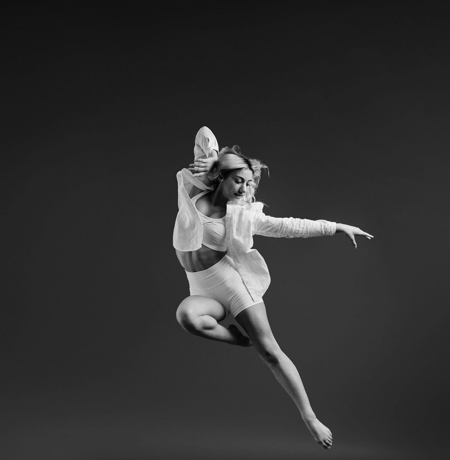 A person elegantly leaps mid-air in a monochromatic setting, wearing a light outfit against a plain dark background, conveying grace and motion.