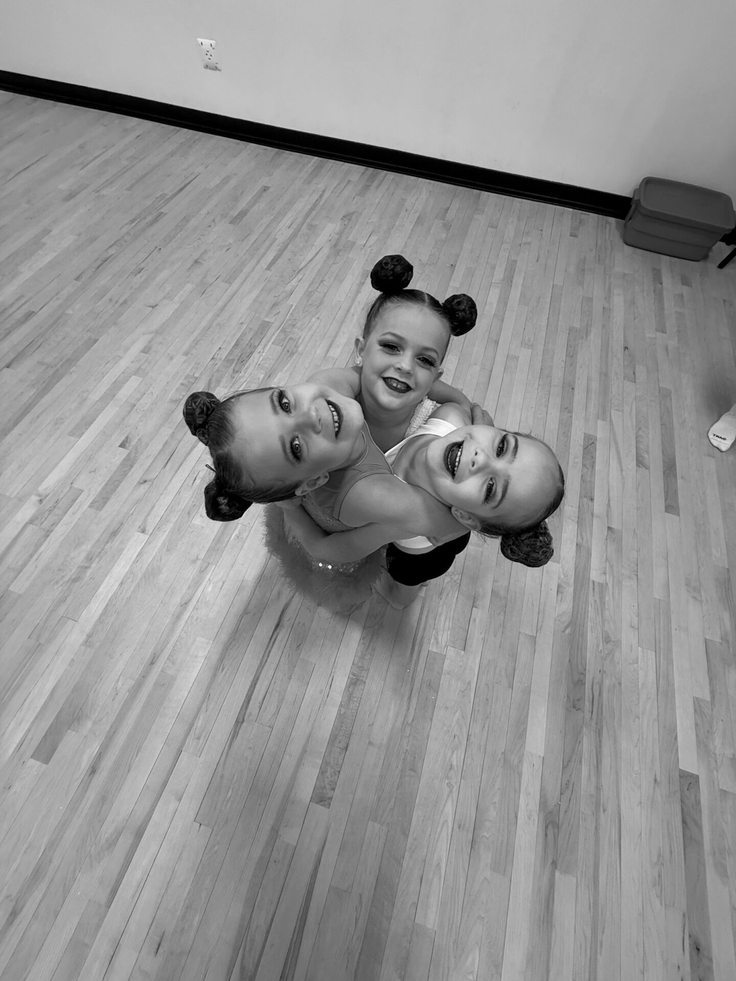 Three smiling children with styled hair, hugging in a dance studio with wooden floors. The image is in black and white.