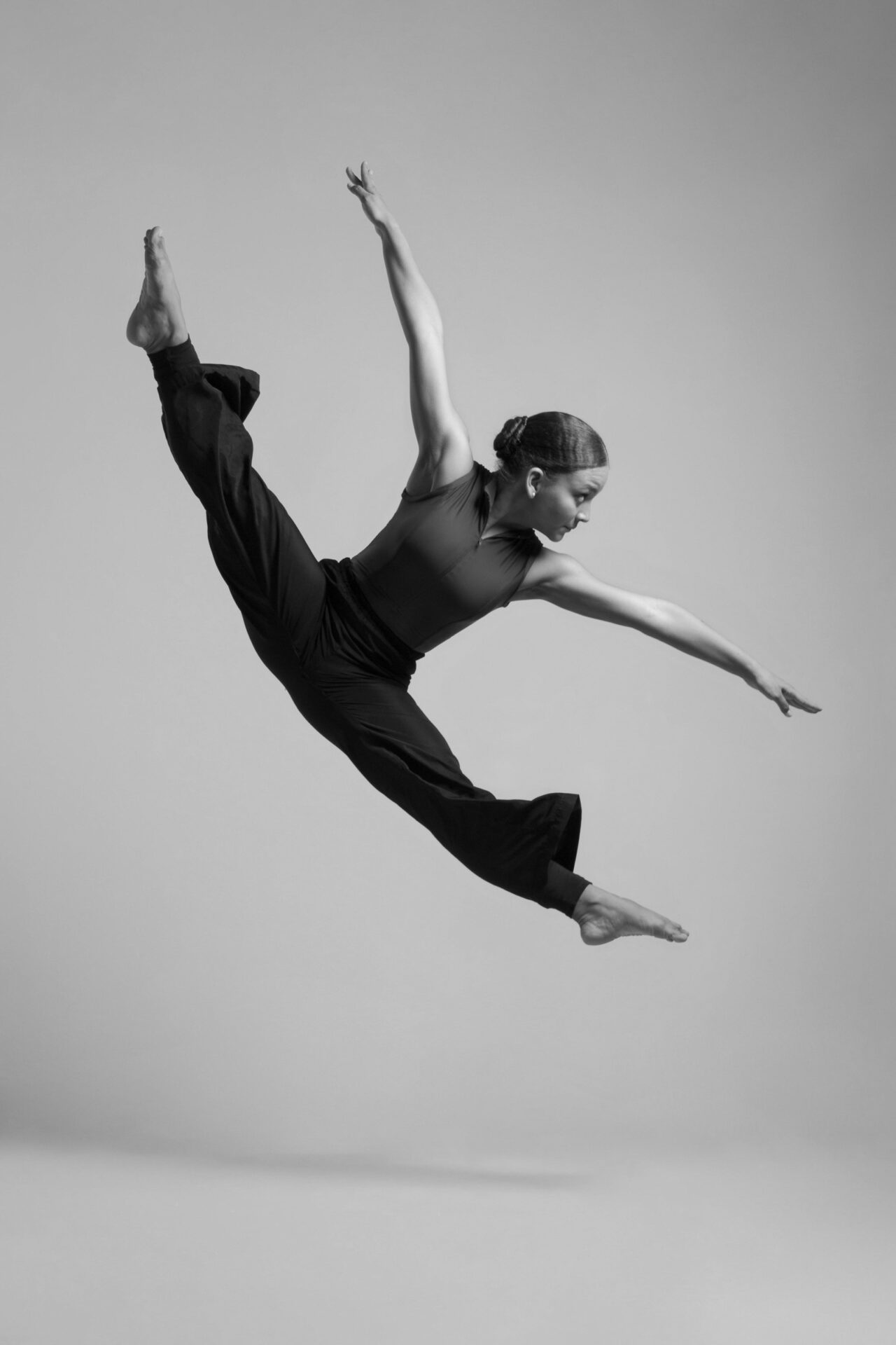 A person gracefully performs an airborne split leap, showcasing flexibility and strength against a plain background in a black and white photograph.