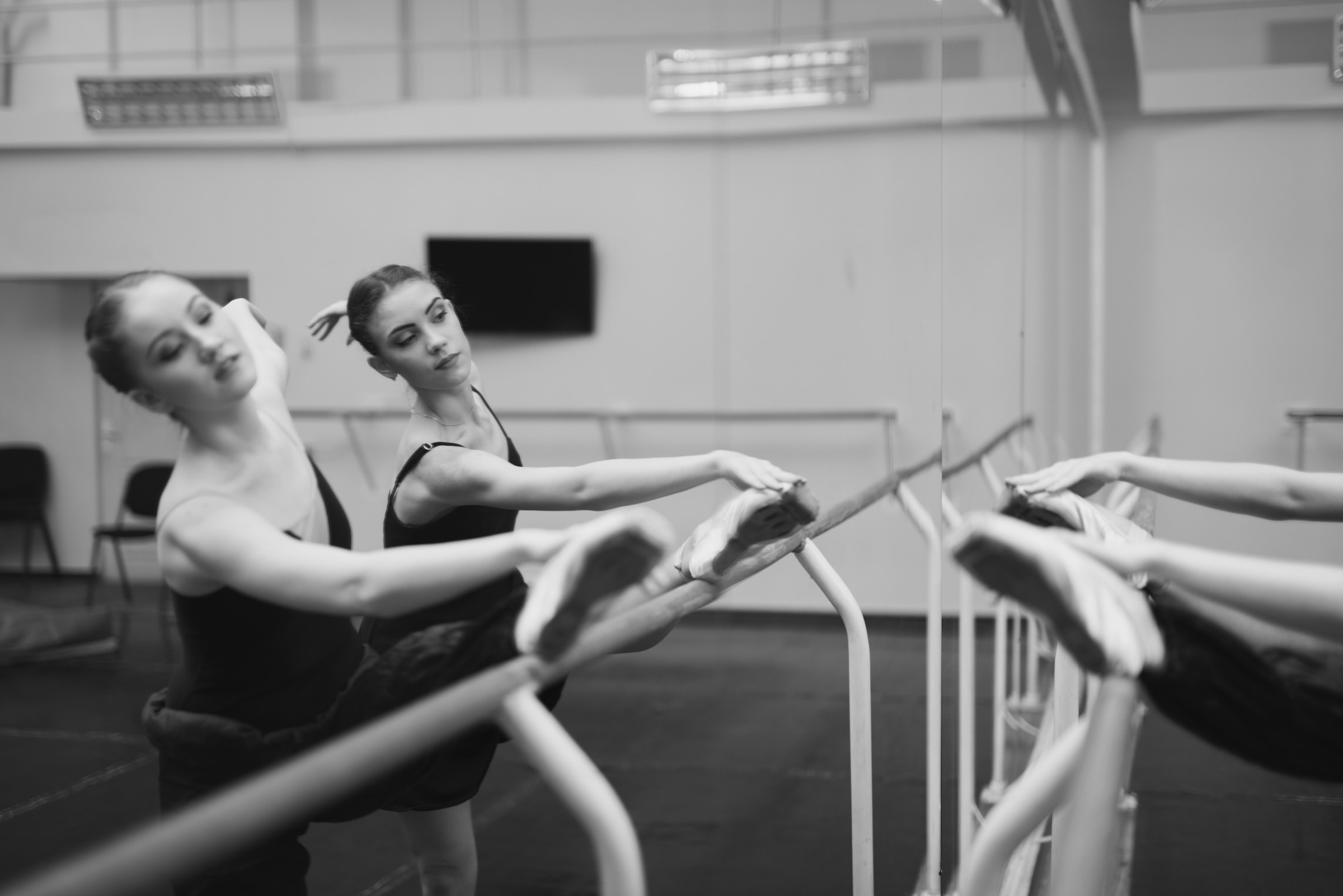 Two persons practice ballet at a barre in a mirrored dance studio, focusing on stretching and form, creating a calm and elegant atmosphere.