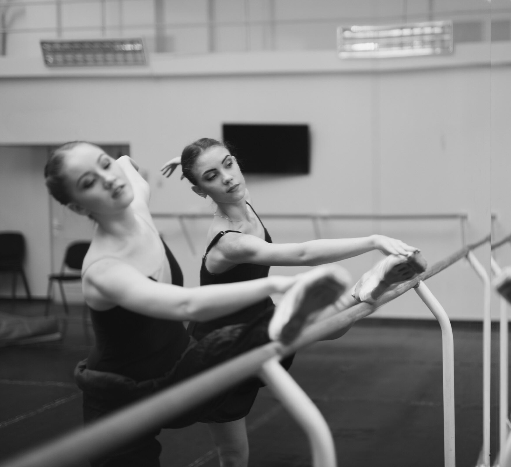 Two people practicing ballet at a barre, stretching with their legs raised, reflected in large mirrors inside a dance studio.