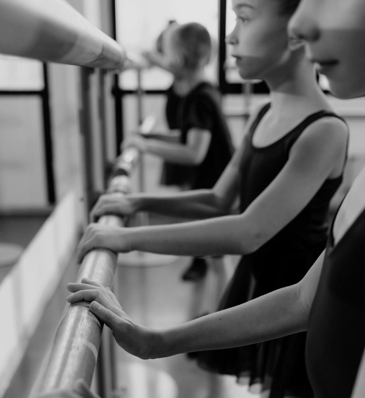 Young dancers practice at a barre in a ballet studio, focusing on alignment and technique. Black and white image emphasizes concentration.
