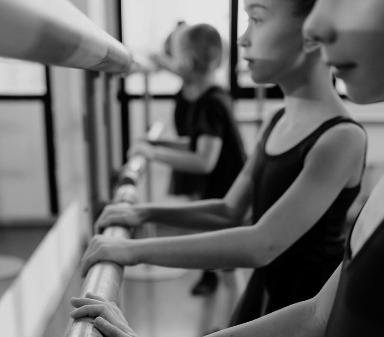 Children in black leotards practice ballet at a barre, focused and poised in a bright studio setting. Black and white photograph.