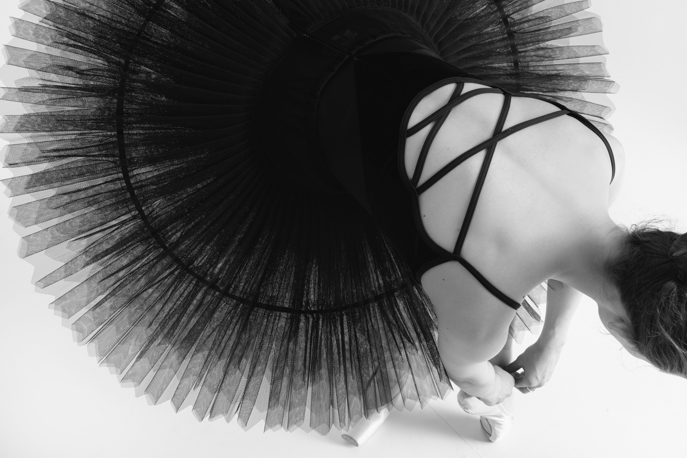 A person in a black tutu adjusts ballet shoes, captured from above in a monochrome setting, highlighting grace and poise.