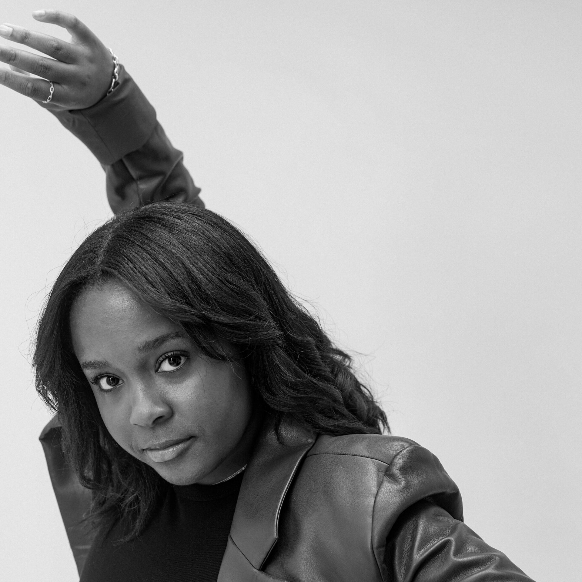 Black and white portrait of a person posing with a raised arm, wearing a leather jacket and textured hairstyle, against a plain backdrop.