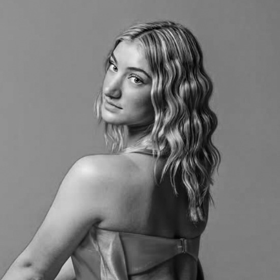 A person with wavy hair looks over their shoulder, wearing a strapless dress, in a studio setting with a plain background.