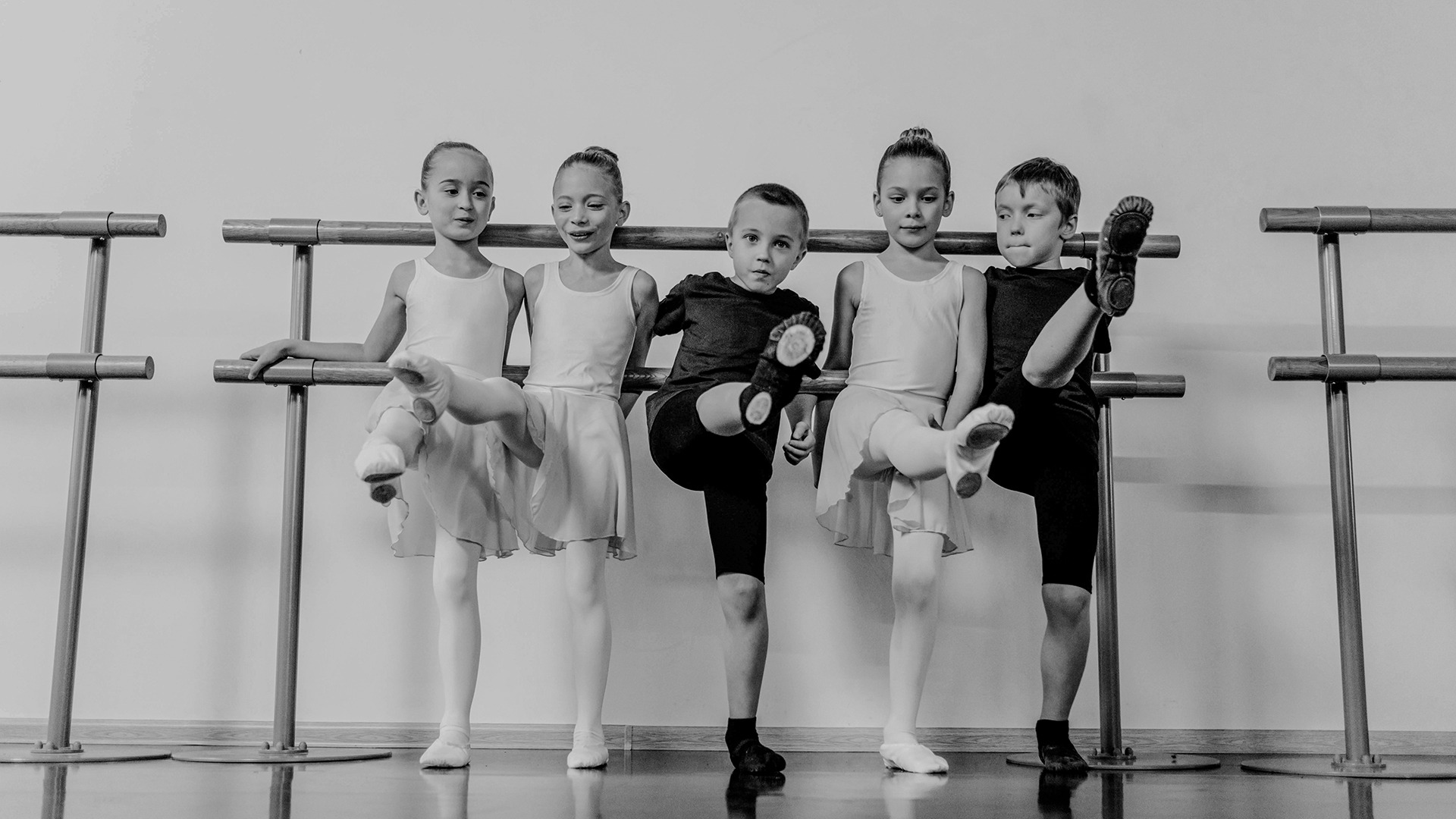 Five children practice ballet at a barre, lifting one leg each, dressed in dance attire in a studio.