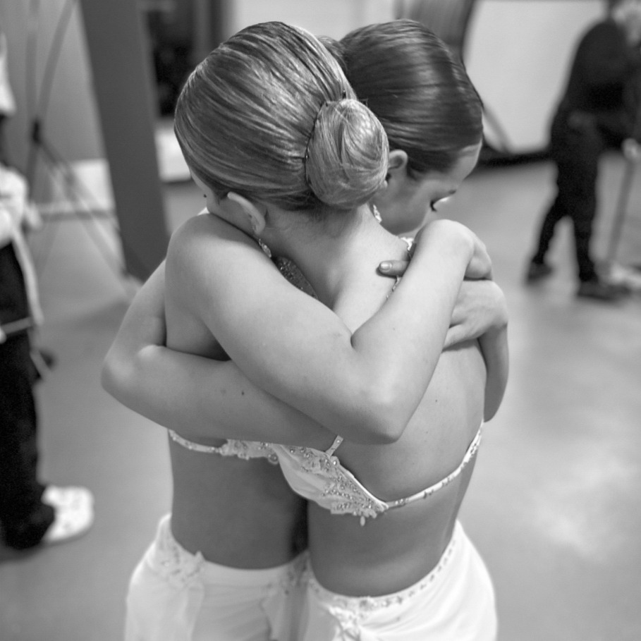 Two people in elegant outfits embrace backstage. Others stand nearby, abstract decor in background; a moment of intimacy and support captured candidly.
