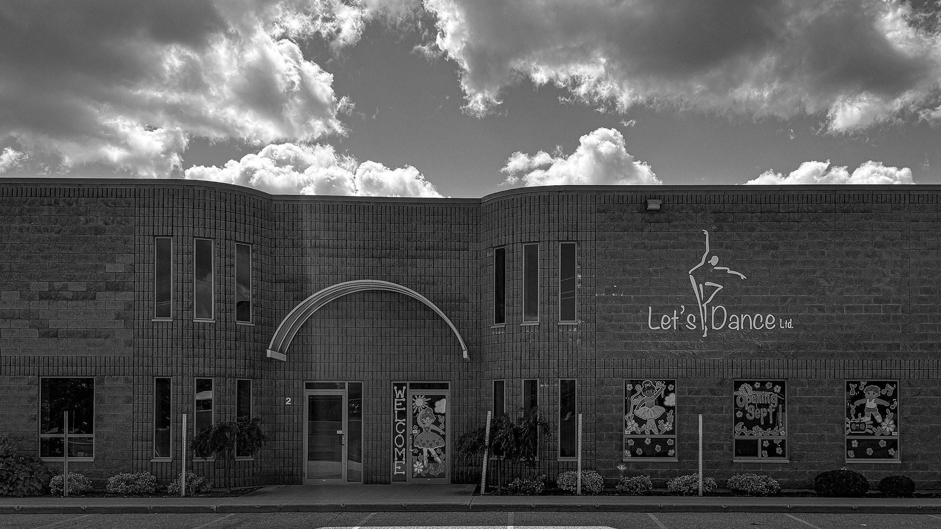 A dance studio with a modern brick facade and welcoming decorations is under a cloudy sky. Sign reads "Let's Dance Ltd."
