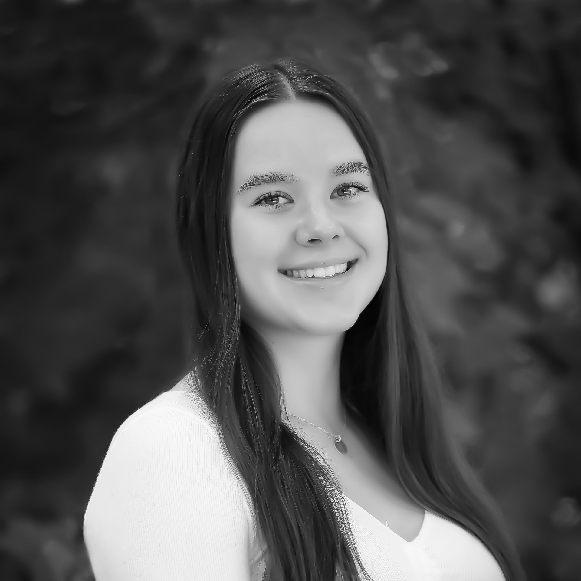 Smiling person with long hair in a black and white photo, standing against a softly blurred natural background.