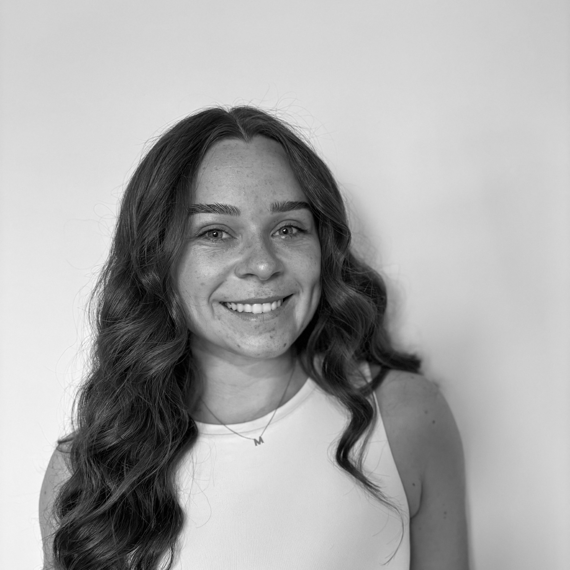 A person with long hair smiles at the camera, wearing a necklace with an "M" pendant, against a plain background. Black and white photo.