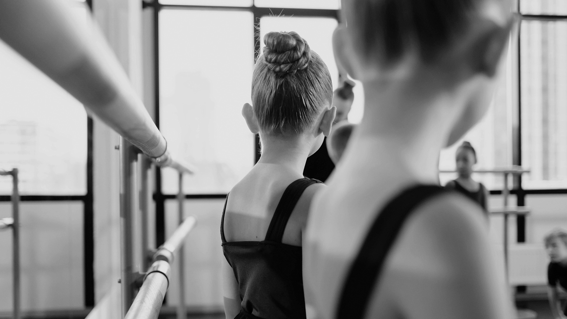 Ballet class scene with children in focus, practicing at a barre in front of large studio windows. Black and white photograph.