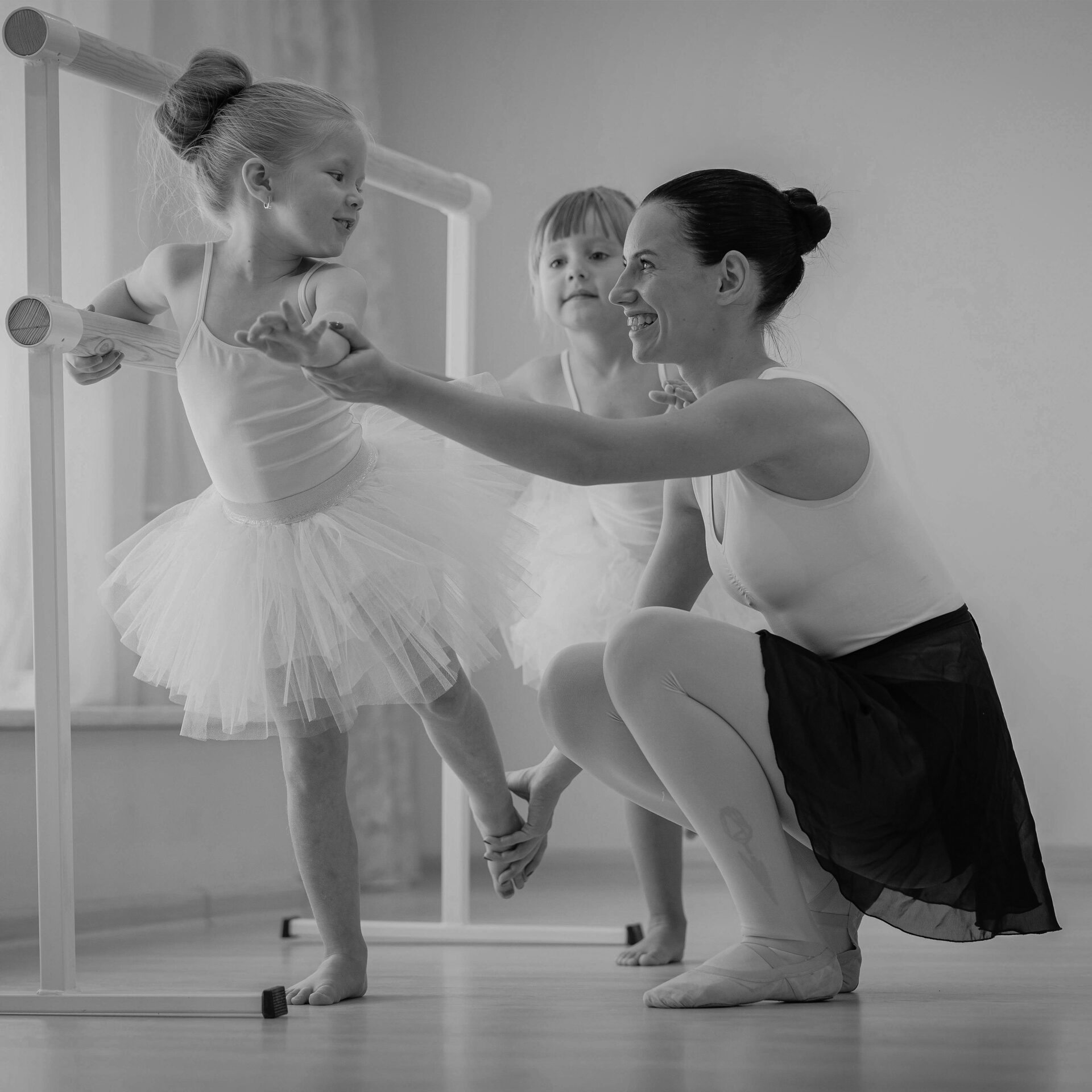 A ballet instructor assists two young students at the barre in a dance studio, focusing on posture and balance.