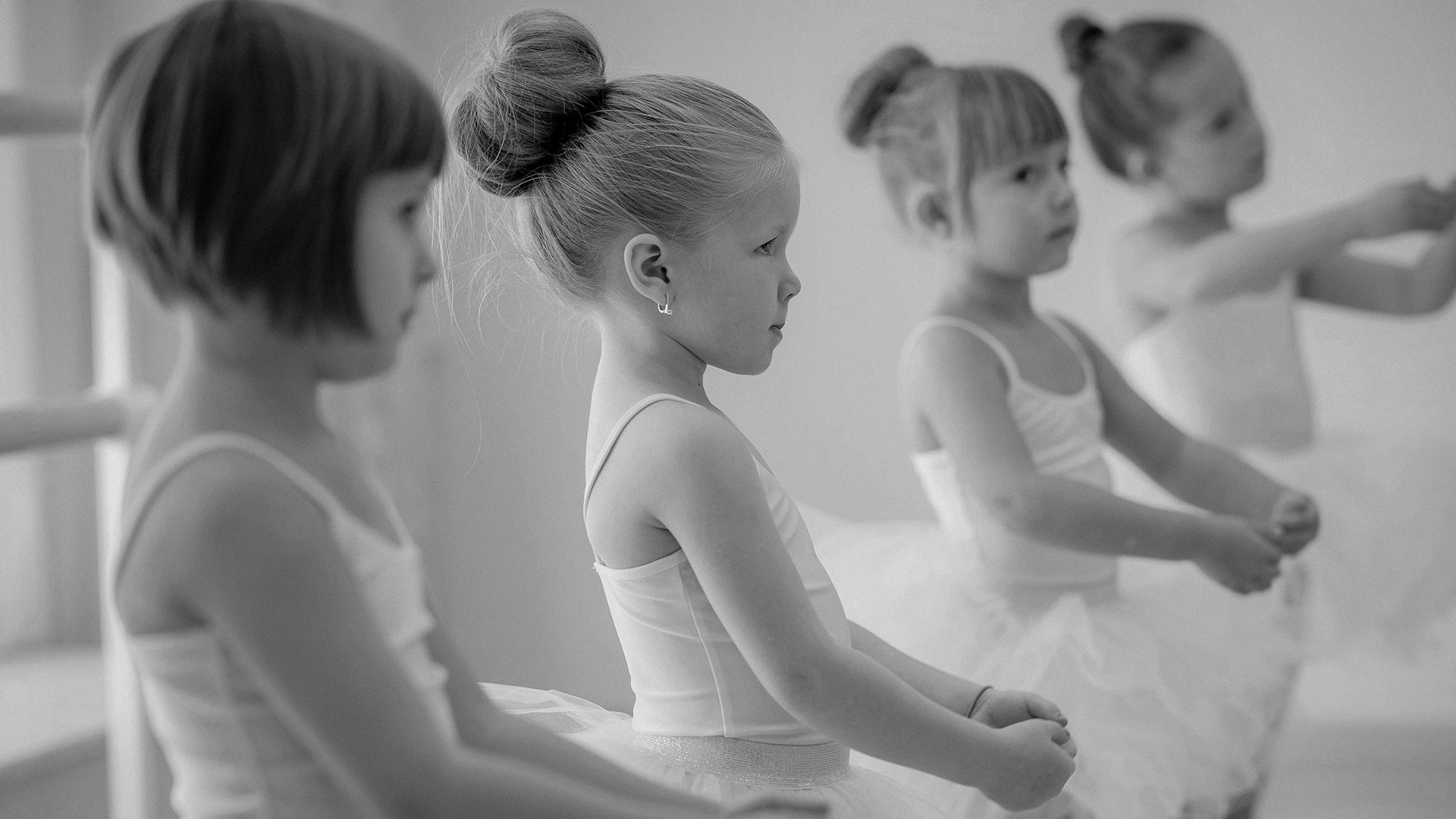 Four young ballet dancers stand in a line, wearing tutus, focused on their practice in a bright dance studio.