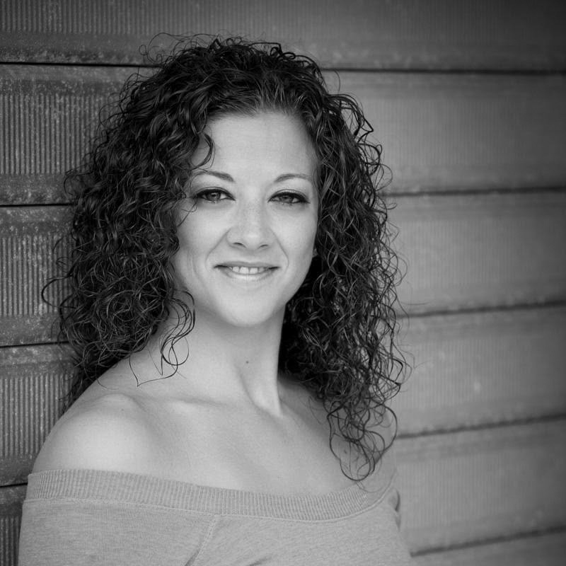 A person with curly hair smiles while leaning against a textured wall. The image is black and white, creating a classic portrait feel.